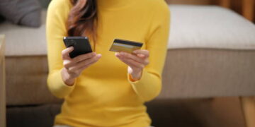 Young happy woman using mobile cell phone credit bank card shopping online booking tour sit on sofa stay home flat rest relax spend free spare time in living room indoors.
