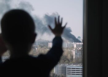 Silhouette of small child. Kid stands with his palms on the window and looks at the fire in city. Black smoke rises into the sky above high-rise buildings. Boy is worried. Child safety.