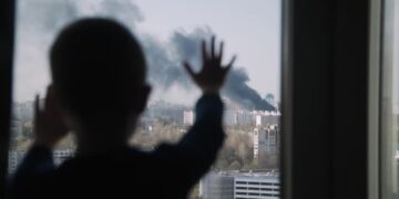Silhouette of small child. Kid stands with his palms on the window and looks at the fire in city. Black smoke rises into the sky above high-rise buildings. Boy is worried. Child safety.