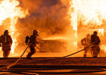4 firefighters battle a roaring blaze.  2 fire hoses shooting water.