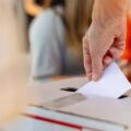 Close-up of hand placing ballot into voting box at election. Ballot and voting box symbolize democracy and elections. Hand placing ballot in election voting box, casting a vote at election