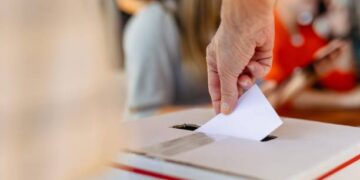 Close-up of hand placing ballot into voting box at election. Ballot and voting box symbolize democracy and elections. Hand placing ballot in election voting box, casting a vote at election