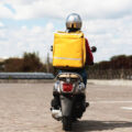 Delivery Service. Courier With Yellow Backpack Riding Scooter Back To Camera In Urban Area Delivering Food In City. Copy Space