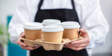 Waiter barista holding and serving take out a paper disposable cup of hot coffee in cafe
