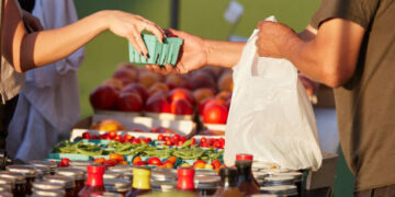 Farmer at market hands purchase to customer over colorful blend of organic produce