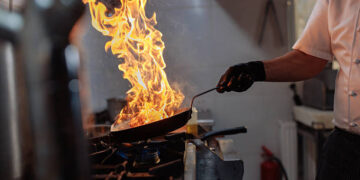 Chef flambeing food in a pan over high heat in a commercial kitchen, creating flames and smoke