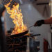 Chef flambeing food in a pan over high heat in a commercial kitchen, creating flames and smoke