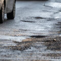 Close-up of potholes on a wet urban road with a passing vehicle, illustrating damaged infrastructure, poor road conditions, traffic safety risks, and the need for road maintenance in city environments.