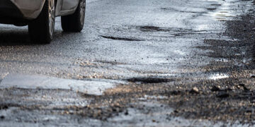 Close-up of potholes on a wet urban road with a passing vehicle, illustrating damaged infrastructure, poor road conditions, traffic safety risks, and the need for road maintenance in city environments.