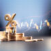 Percentage sign on top of coin stacks before blue financial graph. Horizontal composition with copy space. Business and finance concept.
