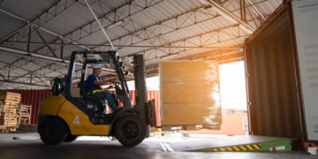 Warehouse worker driver in uniform delivery and loading pallet product by forklift.